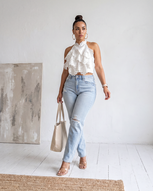 Front image full-body view of the Madison Distressed Straight Denim Jean in a light wash featuring distressed knees and a straight-leg fit, styled with a white ruffled top and a tote bag.